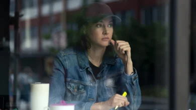"Woman in denim jacket and baseball cap sits at a table inside a cafe, looking thoughtful while holding a yellow highlighter, with a mug and papers in front of her, city street visible through the window."
