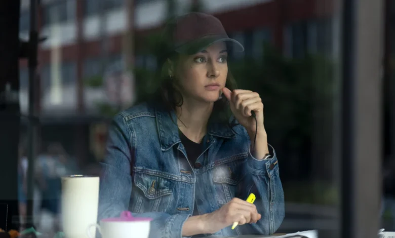 "Woman in denim jacket and baseball cap sits at a table inside a cafe, looking thoughtful while holding a yellow highlighter, with a mug and papers in front of her, city street visible through the window."